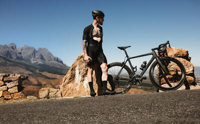 Low angle shot of male road biker standing on the mountain road with his bike. Profi-Radfahrer macht Pause beim Training auf der Bergstraße. - JLPSF18369