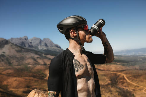 Männlicher Radfahrer, der Wasser aus einer Flasche trinkt. Sportler, der eine Pause macht und Wasser während einer Fahrradtour auf einer Bergstraße trinkt. - JLPSF18373