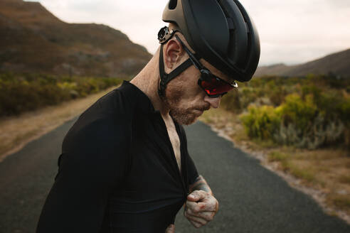 Nahaufnahme eines männlichen Radfahrers mit Helm und Sonnenbrille, der sein Hemd zuzieht. Ein Profi-Radfahrer macht sich auf einer Landstraße für das Radtraining bereit. - JLPSF18380
