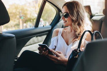 Geschäftsfrau mit Sonnenbrille, die auf dem Rücksitz eines Autos sitzt, aus dem Fenster schaut und lächelt. - JLPSF18388
