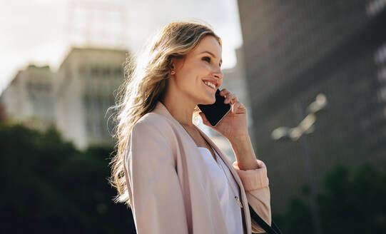 Geschäftsfrau, die ein Handy benutzt und draußen spazieren geht. Lächelnde weibliche Führungskraft, die in der Stadt spazieren geht und einen Anruf tätigt. - JLPSF18392