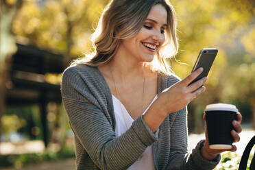 Geschäftsfrau sitzt im Freien und telefoniert. Immobilienmaklerin mit einer Tasse Kaffee in der Hand, die draußen eine Textnachricht auf ihrem Mobiltelefon liest. - JLPSF18397