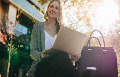 Frau mit Kopfhörern, die draußen sitzt und einen Laptop benutzt. Geschäftsfrau, die an einem sonnigen Tag mit einem Laptop draußen in der Stadt sitzt. - JLPSF18399