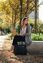 Lächelnde junge Frau sitzt im Freien mit Laptop. Frau schaut weg und lächelt, während sie draußen in der Stadt sitzt. - JLPSF18400