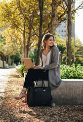 Smiling young woman sitting outdoors with laptop. Woman looking away and smiling while sitting outside in the city. - JLPSF18400