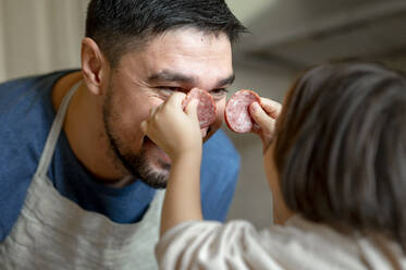 Son holding pepperoni over fathers face in kitchen at home - ANAF00286