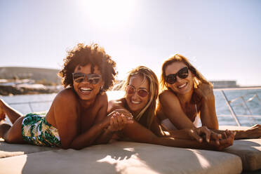 Eine gemischtrassige Gruppe von Frauen mit Sonnenbrillen liegt an einem Sommertag auf dem Bug einer Yacht. Drei junge Frauen nehmen ein Sonnenbad auf einer privaten Yacht. - JLPSF18408