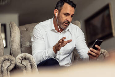 Älterer Geschäftsmann mit Kopfhörern, der mit seinem Handy telefoniert. Ein Mann auf Geschäftsreise sitzt im Hotelzimmer und telefoniert. - JLPSF18418