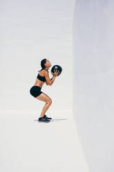 Female athlete doing squats holding a medicine ball. Side view of a woman in fitness wear doing workout using a medicine ball. - JLPSF18490