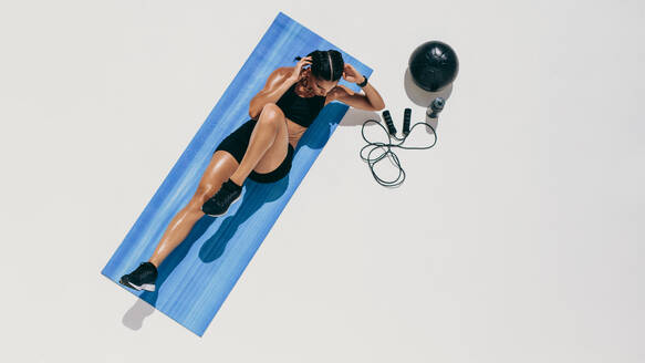 Draufsicht auf eine Sportlerin, die Bauchübungen macht: Frau in Fitnesskleidung, die mit einem Medizinball und einem Springseil an der Seite Bauchpressen macht. - JLPSF18497