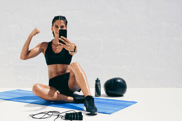Woman in fitness wear sitting on yoga mat taking a selfie showing her biceps. Fitness woman looking at her phone during workout with a medicine ball and skipping rope by her side. - JLPSF18499