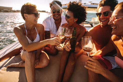 Eine Gruppe junger Männer und Frauen sitzt auf dem Deck einer Jacht und stößt mit Getränken an. Junge Leute feiern auf einem Boot mit Getränken. - JLPSF18521