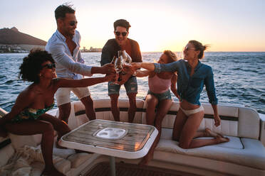 Group of diverse men and women sitting at the back of the boat and toasting drinks. Young people partying on a boat with drinks at sunset. - JLPSF18549