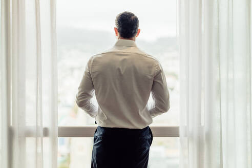 Rückansicht eines Mannes in formeller Kleidung, der am Fenster eines Hotelzimmers steht und nach draußen blickt. Geschäftsmann, der vom Fenster eines Hotelzimmers aus die Stadtlandschaft betrachtet. - JLPSF18553