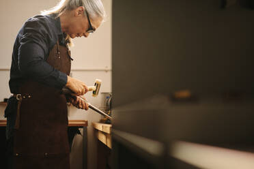 Ältere Frau, die in ihrer Werkstatt mit traditionellen Werkzeugen Schmuck herstellt. Goldschmiedin mit Lederschürze und Brille, die an einer Werkbank mit Hammer und Stahlmaßwerkzeug einen Ring herstellt. - JLPSF18569