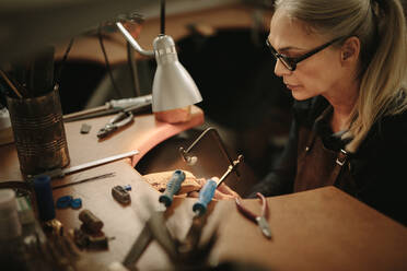 Ein erfahrener Schmuckhersteller schneidet Metall mit einer Bandsäge auf einer Werkbank. Eine professionelle Juwelierin fertigt einen Ring in ihrer Werkstatt an. - JLPSF18570