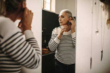 Reflektion einer älteren Frau, die einen Designer-Ohrring beim Juwelier anprobiert. Eine Frau kauft einen Ohrring und schaut in den Spiegel im Juweliergeschäft. - JLPSF18608