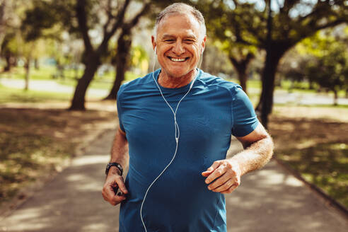 Porträt eines älteren Mannes in Fitnesskleidung, der in einem Park läuft. Nahaufnahme eines lächelnden Mannes, der läuft, während er mit Kopfhörern Musik hört. - JLPSF18612