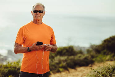 Älterer Mann in Fitnesskleidung, der im Freien steht und ein Mobiltelefon hält. Fitness-Mann mit dunkler Brille, der während des Trainings im Freien steht und ein Mobiltelefon und Kopfhörer hält. - JLPSF18618