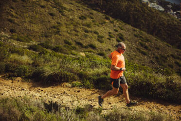Athletischer Mann, der an einem sonnigen Tag einen Hügel hinunterläuft. Blickwinkel eines älteren Mannes in Fitnesskleidung, der auf einem rauen, hügeligen Terrain läuft. - JLPSF18624