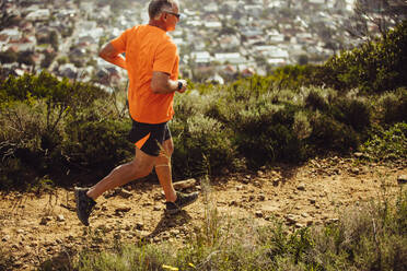 Seitenansicht eines älteren Mannes beim Trailrunning auf einem Hügel. Älterer athletischer Mann, der an einem sonnigen Tag eine raue, hügelige Landschaft hinunterläuft. - JLPSF18625