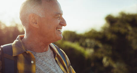Seitenansicht eines lächelnden Mannes, der im Freien steht, mit der Sonne im Hintergrund. Ausgeschnittenes Porträt eines lächelnden älteren Mannes, der wegschaut. - JLPSF18634