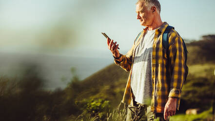 Ein älterer Mann mit Rucksack steht auf einer Anhöhe und schaut auf sein Handy. Ein Mann schaut auf sein Handy nach einer erfolgreichen Wanderung auf einem Berg. - JLPSF18640
