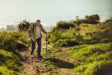Ein Mann geht auf einem hügeligen Pfad und hält Trekkingstöcke. Ein abenteuerlustiger Mann auf einer Trekkingtour in einem hügeligen Gelände mit dem Meer im Hintergrund. - JLPSF18641