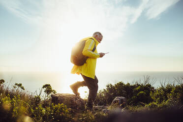 Side view of a man wearing backpack walking on a rocky hill. Senior man hiking on a hill looking at his mobile phone with sun in the background. - JLPSF18646