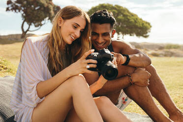 Close up of a tourist couple on vacation sitting outdoors looking at the photos in a digital camera. Smiling man and woman on holiday sitting in a park checking their photos taken in a dslr camera. - JLPSF18670