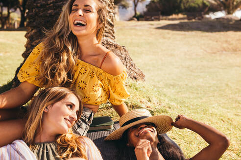 Drei fröhliche Frauen, die in einem Park sitzen und sich amüsieren. Nahaufnahme von drei Freundinnen, die an einem sonnigen Tag in einem Park auf dem Rasen sitzen und lachen und sich entspannen. - JLPSF18674