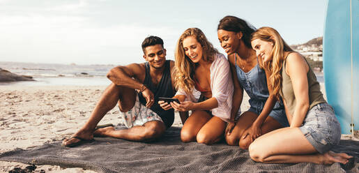 Lächelnde Frau, die ein Selfie mit ihren Freunden am Strand macht, mit einem Surfbrett im Hintergrund. Mann und Frauen im Urlaub sitzen zusammen am Strand und schauen auf ein Handy. - JLPSF18679