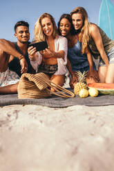 Ebenerdige Aufnahme einer Gruppe von Freunden, die am Strand sitzen und Spaß daran haben, ein Selfie mit dem Handy zu machen. Fröhliche Freunde, die bei einem Picknick am Strand sitzen und lächeln und Gesichter machen, während sie ein Selfie machen. - JLPSF18680