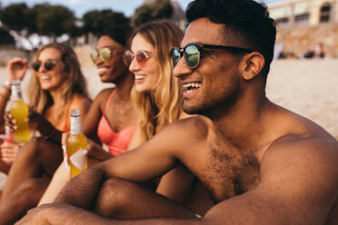 Nahaufnahme einer Gruppe von Freunden im Urlaub, die am Strand sitzen und Sonnenbrillen tragen. Ein lächelnder Mann sitzt mit seinen Freundinnen am Strand und genießt einen Drink. - JLPSF18684