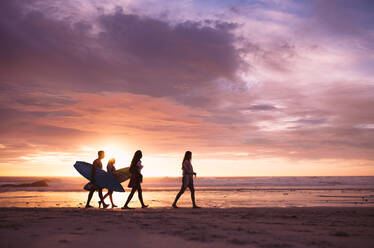 Silhouette einer Gruppe von Freunden, die in der Abenddämmerung am Strand spazieren gehen. Freunde im Urlaub, die bei Sonnenuntergang mit Surfbrettern am Strand spazieren gehen. - JLPSF18692