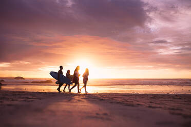 Menschen, die bei Sonnenuntergang am Strand spazieren gehen. Ein Paar im Urlaub, das mit Surfbrettern am Strand spazieren geht. - JLPSF18693