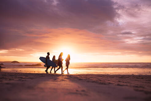 Menschen, die bei Sonnenuntergang am Strand spazieren gehen. Ein Paar im Urlaub, das mit Surfbrettern am Strand spazieren geht. - JLPSF18693