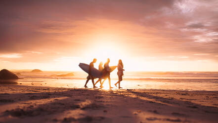 Silhouette eines Menschen, der in der Abenddämmerung am Strand spazieren geht. Männer und Frauen, die bei Sonnenuntergang am Strand spazieren gehen und Surfbretter tragen. - JLPSF18694