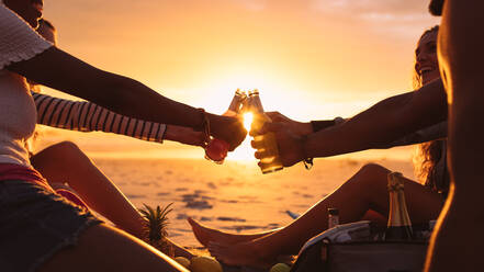 Beschnittene Aufnahme von Freunden, die bei Sonnenuntergang mit Essen und Getränken am Meer sitzen. Fröhliche Freunde im Urlaub, die am Strand mit der Sonne im Hintergrund auf Getränke anstoßen. - JLPSF18696
