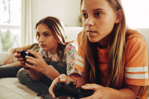 Nahaufnahme von zwei Mädchen im Teenageralter, die ein Videospiel spielen und Joysticks halten. Die Mädchen sitzen zu Hause auf der Couch und spielen mit großem Interesse ein Videospiel. - JLPSF18704