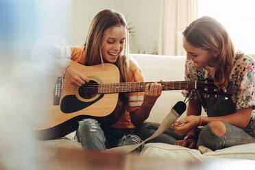 Fröhliche junge Mädchen, die zu Hause Gitarre spielen. Mädchen spielt Musik auf der Gitarre, während ihre Freunde zuschauen. - JLPSF18706