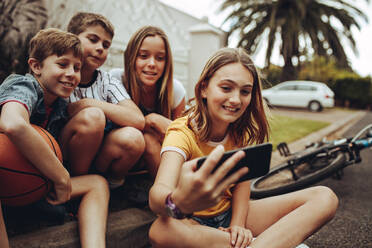 Fröhliche Kinder machen ein Selfie am Straßenrand. Ein Mädchen macht mit ihrem Handy ein Selfie mit ihren Freunden im Freien. - JLPSF18717