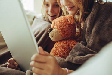 Cropped shot of two girls looking at a tablet pc sitting at home. Close up of two girls sitting at home covered in a blanket and watching movie on a tablet computer. - JLPSF18718