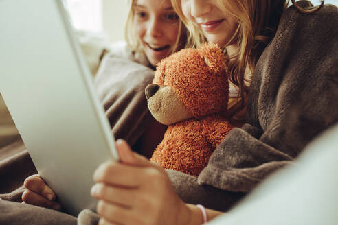 Nahaufnahme von zwei Mädchen, die zu Hause in eine Decke gehüllt sitzen und sich einen Film auf einem Tablet-Computer ansehen. - JLPSF18718