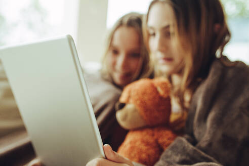 Zwei in eine warme Decke gehüllte Mädchen sehen sich zu Hause einen Film auf einem Tablet-PC an. Ein Mädchen sitzt mit ihrer Schwester zu Hause und hält einen Tablet-Computer und einen Teddybären. - JLPSF18719