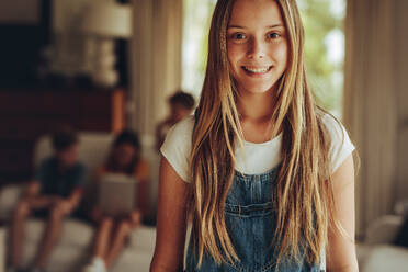 Nahaufnahme eines lächelnden Mädchens mit goldbraunem Haar. Fröhliches Mädchen, das zu Hause steht, während im Hintergrund ihre Freunde sitzen. - JLPSF18722