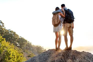 Rear view of hiking couple with backpack standing together on hill top enjoying beautiful landscape. Man and woman outdoors on hiking standing on a rock. - JLPSF18754