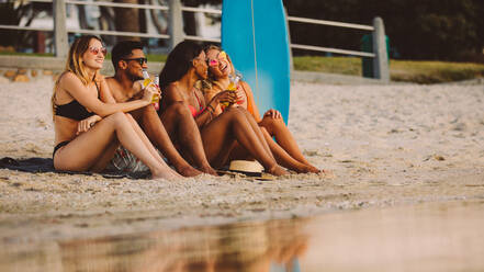 Group of friends sitting together on beach drinking beer. Friends having a great time together on a holiday sitting near the sea. - JLPSF18762