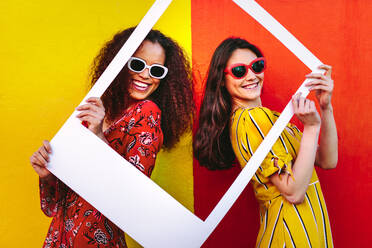 Portrait of beautiful women holding a blank photo frame in hand and smiling. Girls wearing sunglasses standing against red and yellow colored wall. - JLPSF18764