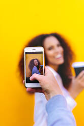 Woman hand with mobile phone taking picture of a female friend holding coffee against yellow background. Female photographing friend with smartphone camera. Focus on female hand holding cellphone. - JLPSF18766